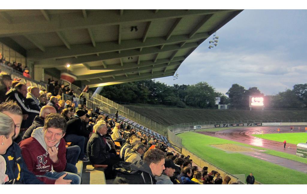 Die mit Zuschauern besetzte Westtribüne des Sportstadions im Bonner Sportpark Nord bei einem abendlichen Heimspiel des Bonner SC (2014)