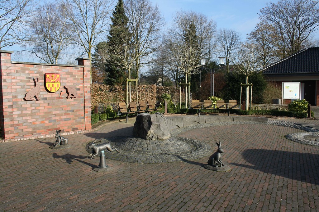Der Brunnen auf dem Dorfplatz in Labbeck mit den drei Hasen-Figuren sowie dem Stadtwappen auf einer Mauer im Hintergrund (2014).