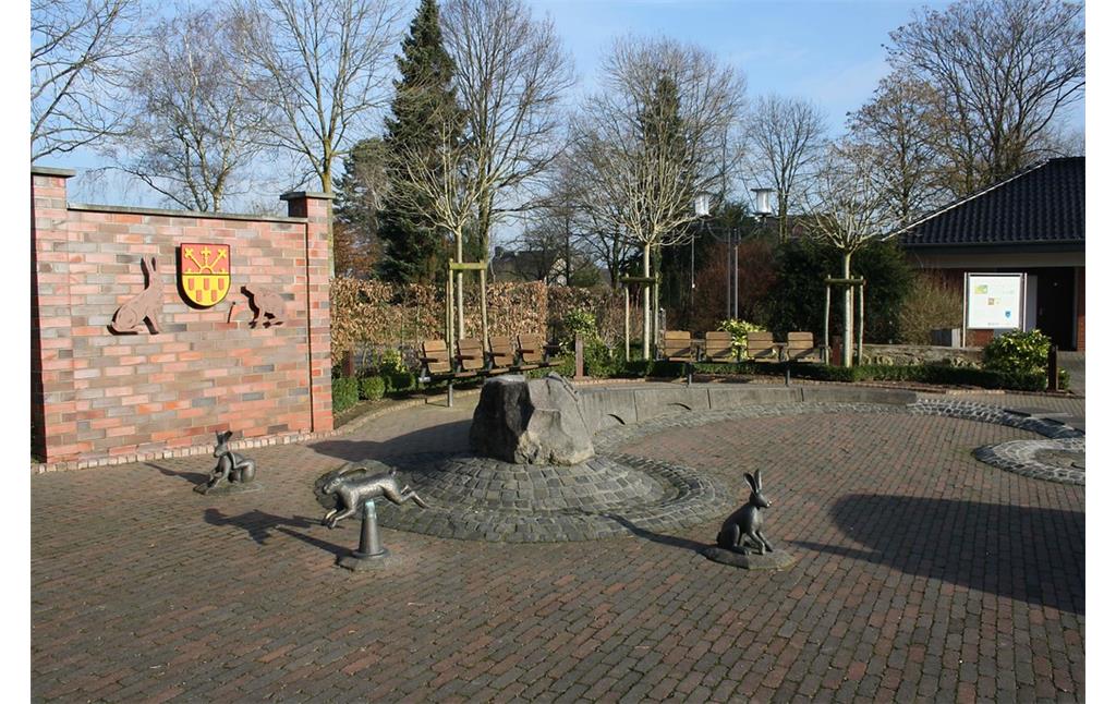 Der Brunnen auf dem Dorfplatz in Labbeck mit den drei Hasen-Figuren sowie dem Stadtwappen auf einer Mauer im Hintergrund (2014).