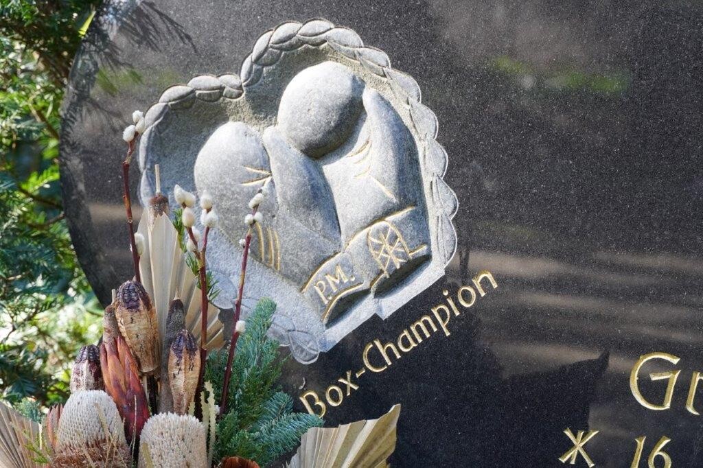 Detail am Grabstein des Kölner Boxers Peter "die Aap" Müller auf dem Kölner Südfriedhof in Zollstock (2023): von einem Siegerkranz umrankte Boxhandschuhe mit den Initialen "P.M." und darunter die Inschrift "Box-Champion".