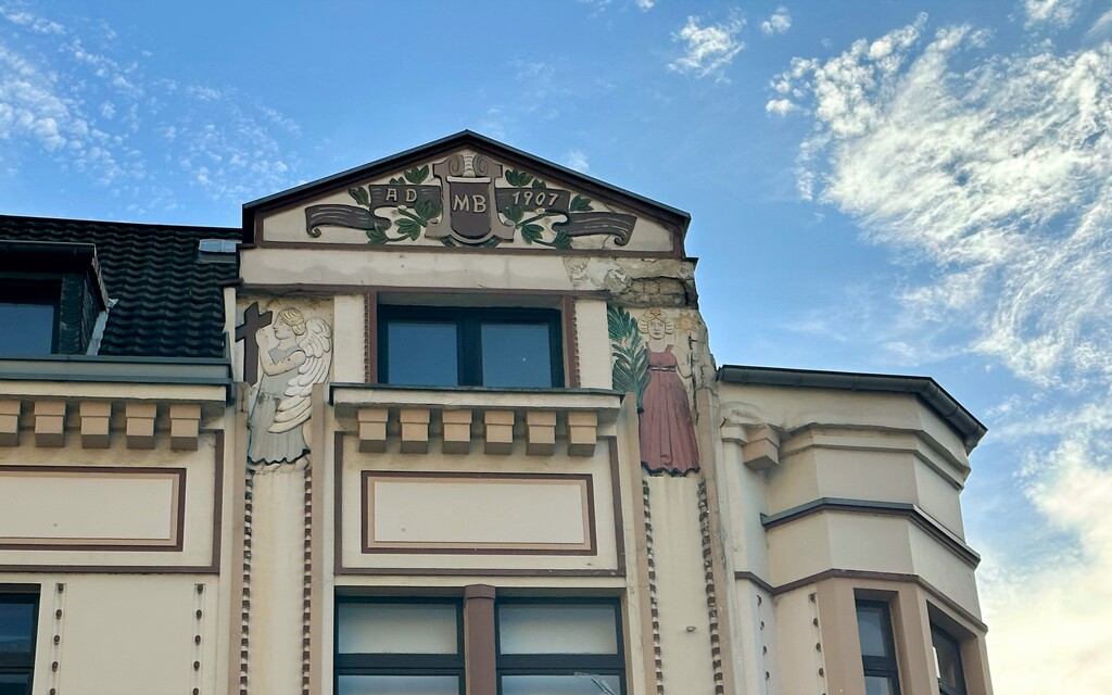 Das Haus Bahnhofstraße Nr. 6a in Porz-Mitte stammt aus dem Jahr 1907. Den Giebel zur Bahnhofstraße hin schmücken die Inschrift AD 1907 sowie die Initialen "MB". Darunter befinden sich links ein Engel mit einem Kreuz in der Hand sowie links eine Frau mit Sonnensymbolik (2023).