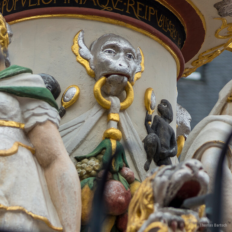 Detail am Petrusbrunnen auf dem Hauptmarkt in Trier (2022): Vier Statuen am Sockel stellen die Kardinaltugenden dar, hinter deren Rücken am Brunnenstock masturbiert ein Affe mit Spiegel.