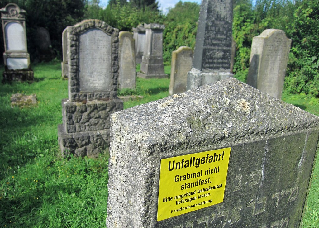 Grabsteine auf dem jüdischen Friedhof in Wevelinghoven. Im Vordergrund befindet sich ein Grabstein mit einem Hinweisschild auf mangelnde Standfestigkeit (2014)