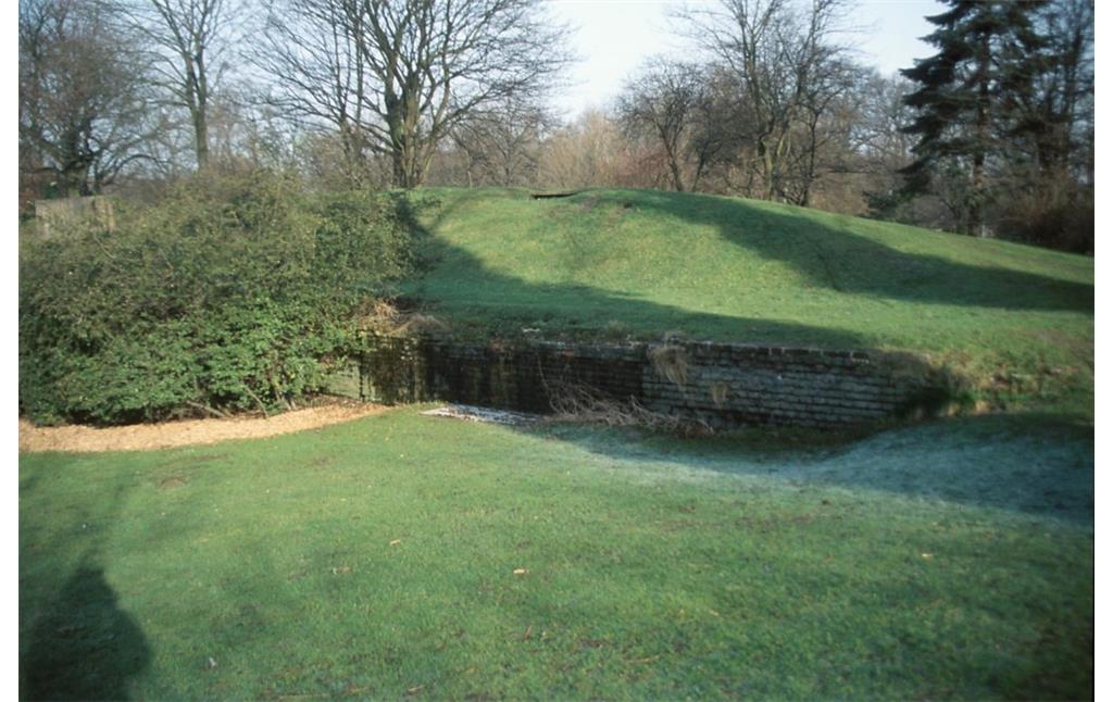 Die Kasematte im Heubergpark von außen. Es handelt sich um einen mit Gras bewachsenen Hügel, an dessen unterem Rand eine Ziegelmauer mit einem vergitterten Bereich zu erkennen ist (2014).
