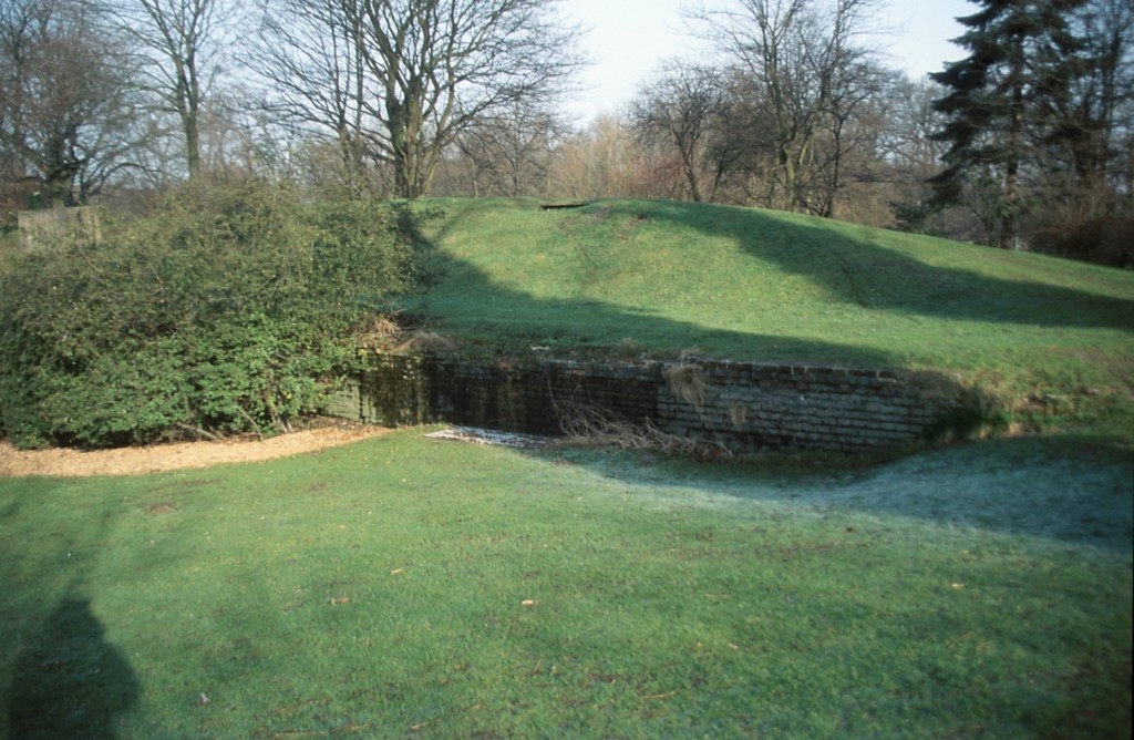 Die Kasematte im Heubergpark von außen. Es handelt sich um einen mit Gras bewachsenen Hügel, an dessen unterem Rand eine Ziegelmauer mit einem vergitterten Bereich zu erkennen ist (2014).